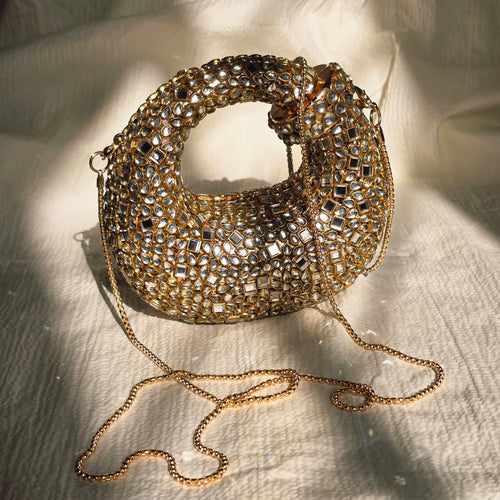 Luxurious gold crescent-shaped clutch bag covered in mirror-style crystals with a detachable gold chain strap, displayed on a textured white fabric background.