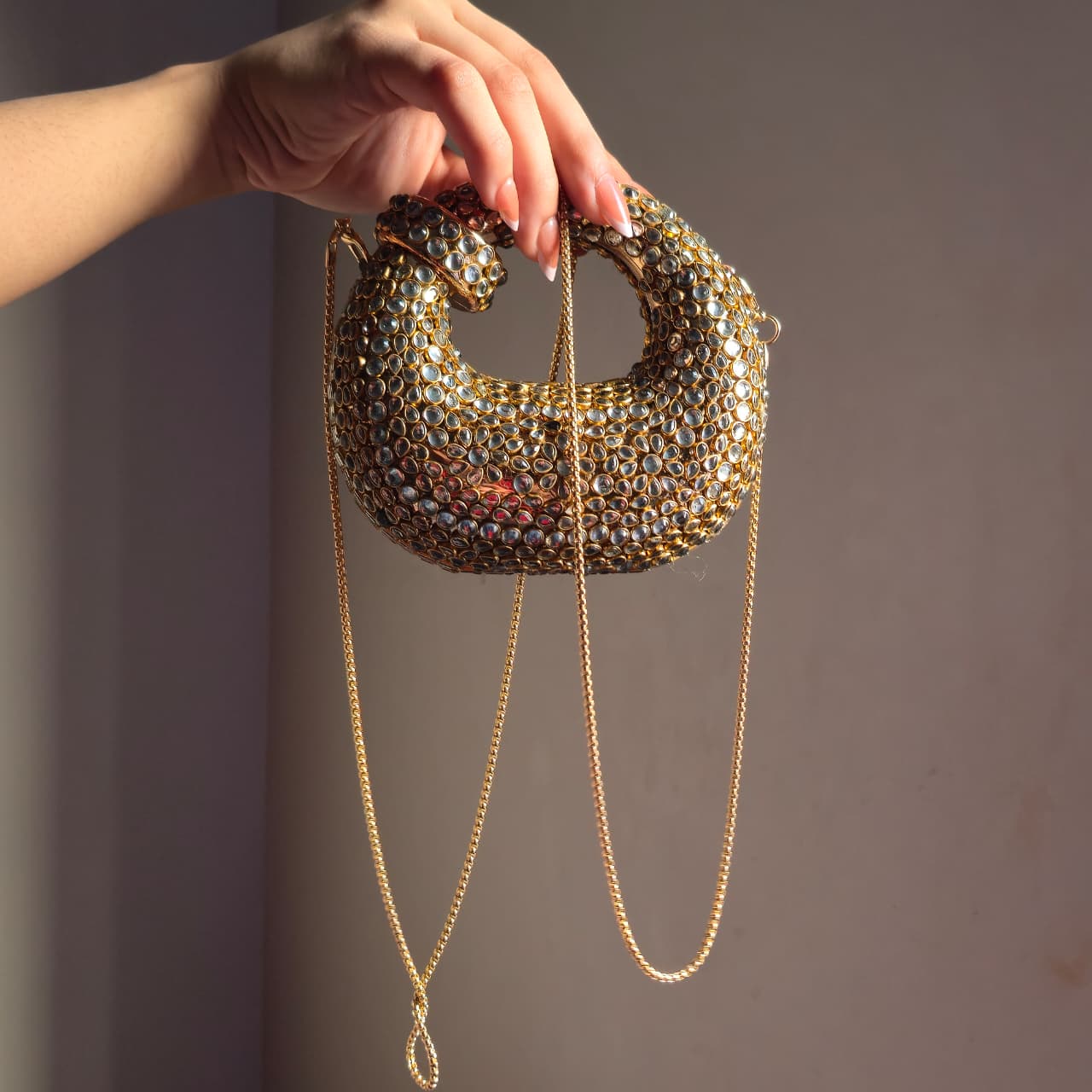 A person holding the gold embellished bag by its handle with the long gold chain strap hanging down to show the length and scale.
