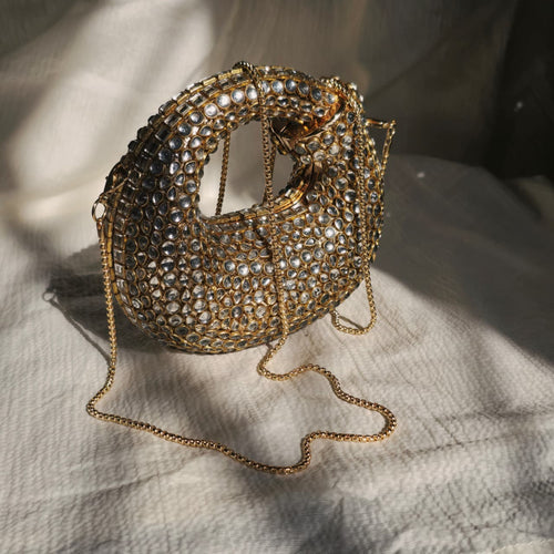 A luxurious gold crescent-shaped evening clutch bag with mirror-crystal embellishments and a gold chain strap on a white fabric background.
