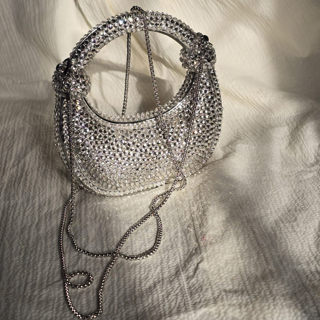 Angled side view of a silver rhinestone hobo-style handbag showing its structured curve and light-reflecting crystal exterior.