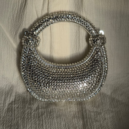 Front view of a crescent-shaped silver evening clutch fully encrusted with shimmering rhinestones on a textured white fabric background.