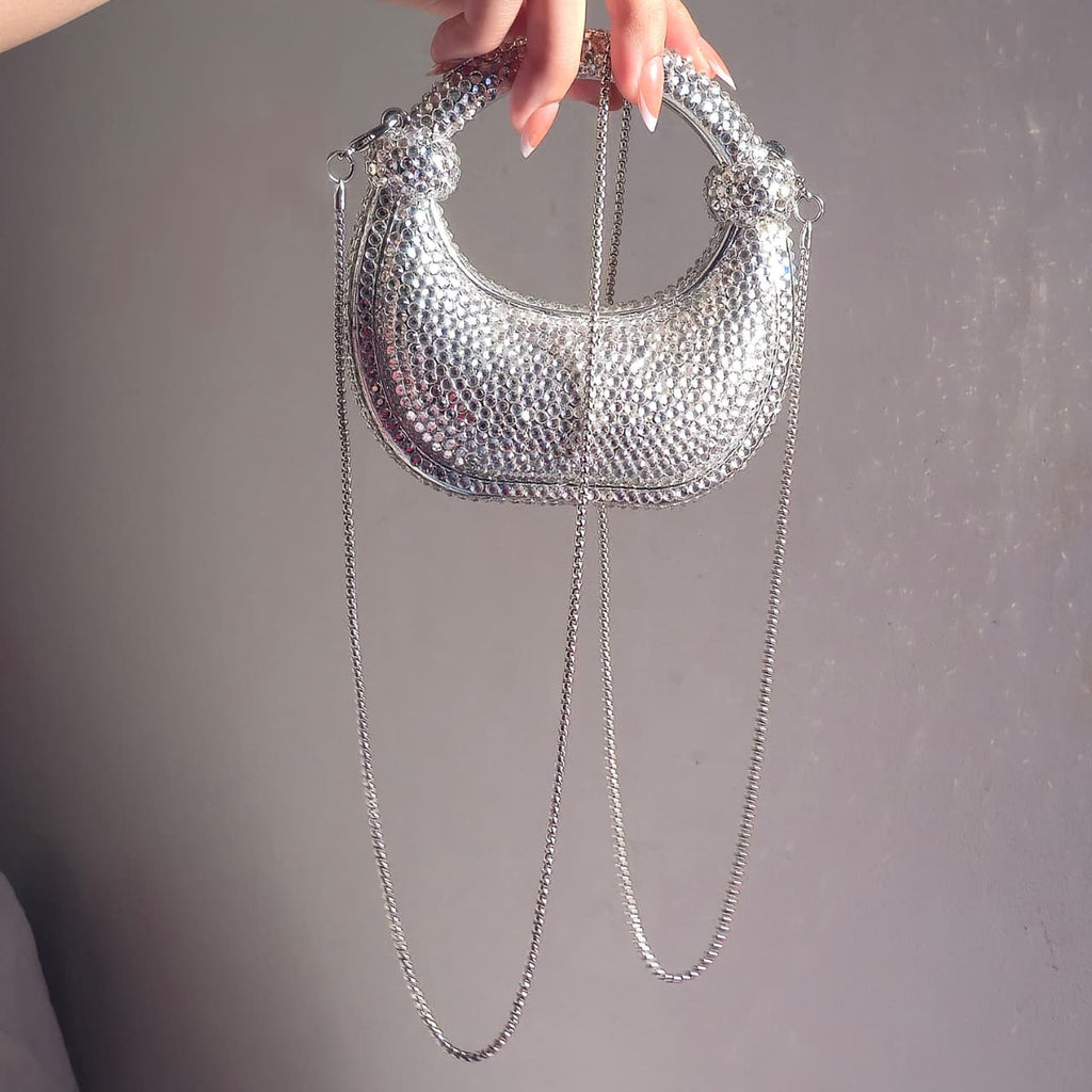 The silver crystal crescent bag being held by its top handle with its detachable silver chain strap visible, showcasing its use as both a handbag and a crossbody.