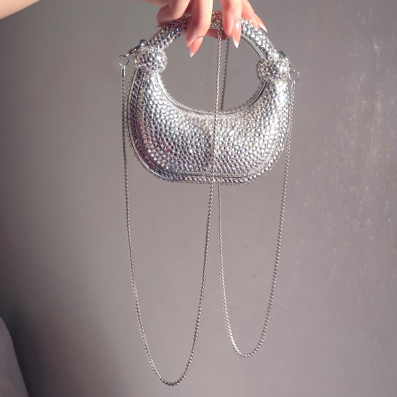 The silver crystal crescent bag being held by its top handle with its detachable silver chain strap visible, showcasing its use as both a handbag and a crossbody.
